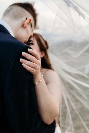 Close-up of bride embracing groom under flowing veil showcasing wedding ring and pearl bracelet.