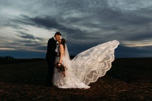 Bride and groom kissing outdoors as wedding veil dramatically flows in the wind under a stormy sky.