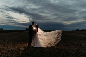 Romantic backlit wedding couple embracing in an open field at dusk with veil billowing behind them.