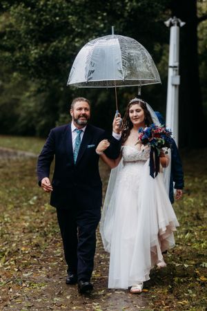 Bride and older gentleman walking outdoors in rain sharing clear dome umbrella, bride holding colorful bouquet.