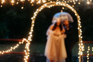 Romantic blurred image of bride and groom standing under glowing arch of fairy lights at dusk.