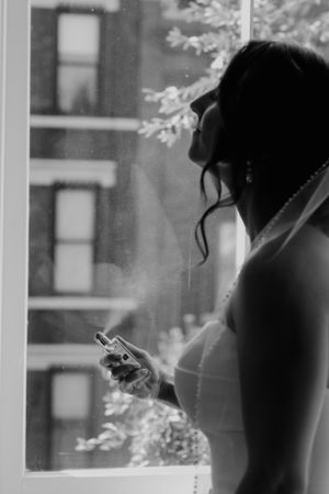 Black and white silhouette of bride in veil holding phone while standing at window with city view.