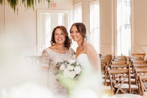 Smiling bride in white gown posing with older woman in beaded dress inside bright elegant venue.