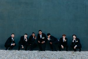 Seven groomsmen in dark suits crouching and laughing against gray wall for fun wedding party photo.