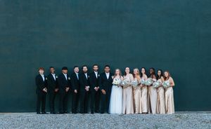 Wedding party poses against teal wall, groomsmen in black suits and bridesmaids in champagne gowns.