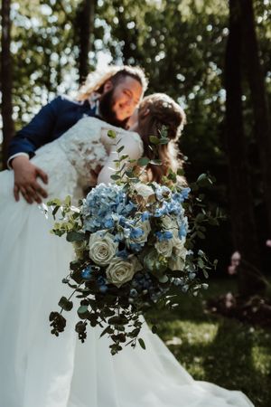 Couple shares a kiss as bride holds blue hydrangea and white rose bouquet in forest setting.