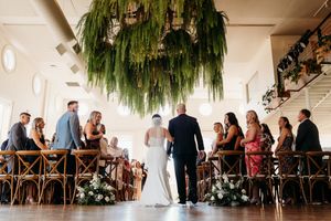 Bride walks down the aisle at an elegant indoor wedding ceremony with lush hanging greenery above.
