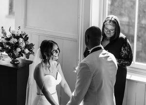 Black and white photo of bride and groom holding hands during intimate indoor wedding ceremony with officiant.