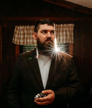 Bearded man in tweed suit holds vintage camera with dramatic lens flare in rustic interior.