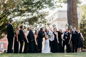 Wedding party in black attire celebrates outdoors under trees as couple shares a romantic kiss.