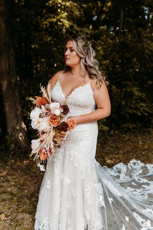 Bride in lace mermaid gown holds autumn bouquet with rust and white blooms in forest setting.