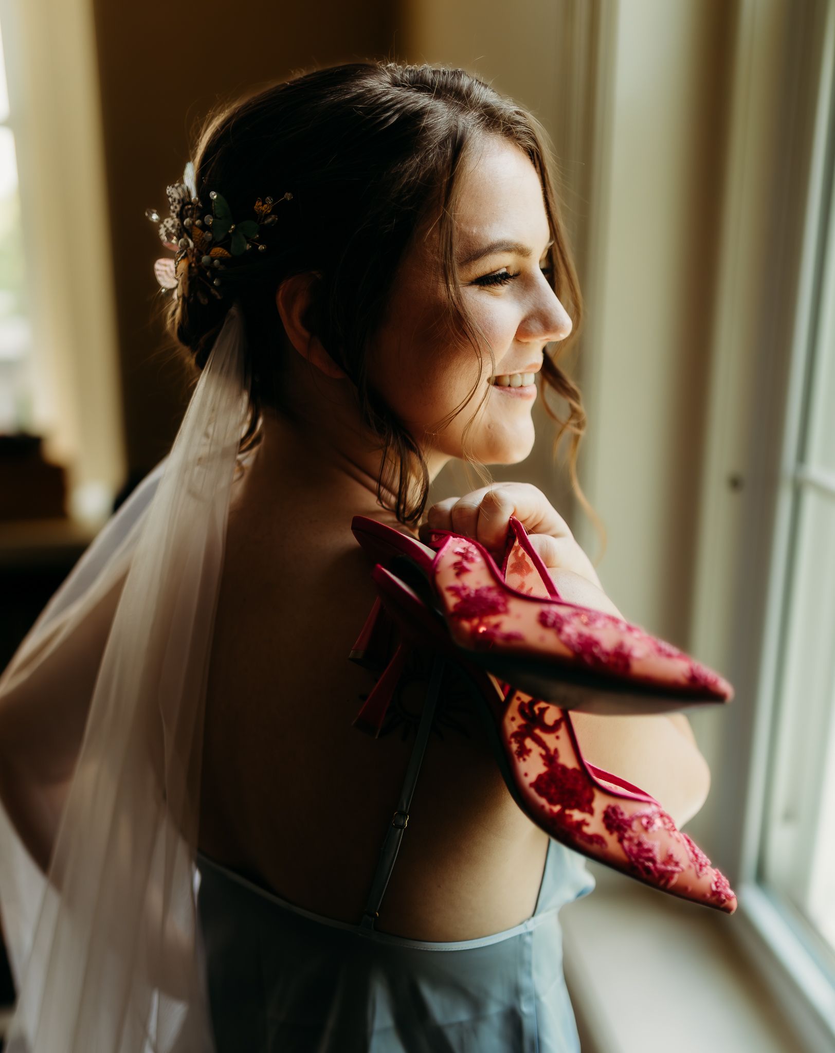 Bride smiles holding pink floral heels over her shoulder, wearing veil and floral hair accessories.