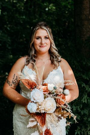 Bride in lace gown holds boho bouquet of terracotta and white flowers before lush greenery.