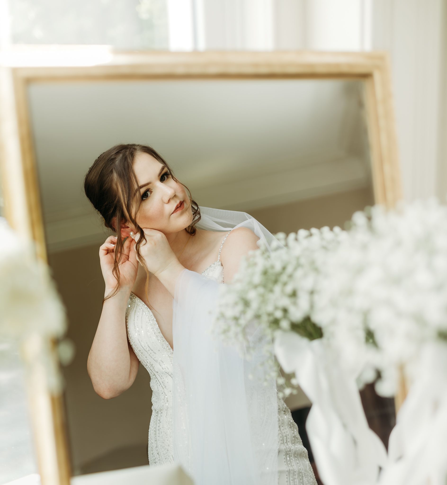 Bride adjusts earring while gazing into gold-framed mirror, wearing white wedding gown and veil.