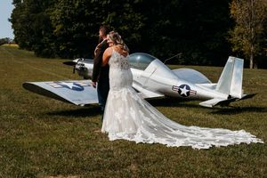 Bride and groom embracing in front of a vintage military airplane on a green grass field.