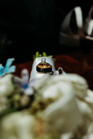 Close-up of gold wedding rings with diamond resting on white floral bouquet in soft dark setting.