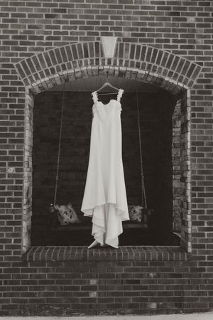 Black and white photo of elegant wedding dress hanging on swing in arched brick wall niche.