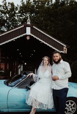 Bride and groom posing beside a classic blue convertible car near a covered bridge.