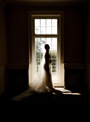 Stunning silhouette of bride in fitted gown and long veil standing before tall Georgian window.