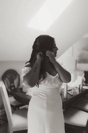 Black and white photo of bride in off-shoulder lace gown putting on earrings while getting ready.