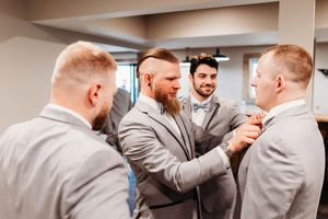 Groomsmen in gray suits help each other with ties while getting ready for wedding ceremony indoors.