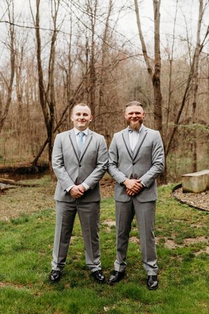 Two groomsmen in matching gray suits smile outdoors at a wedding.