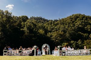 Outdoor wedding ceremony on green lawn with white chairs, floral arch, and lush forested hills under blue sky.