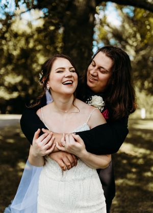 Groom embraces laughing bride from behind outdoors, both sharing a joyful moment together.