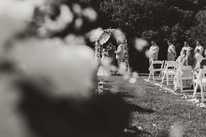 Black and white photo of outdoor wedding ceremony with floral arch and guests seated on lawn.