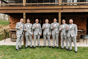 Seven groomsmen in gray suits pose together outside a rustic wooden venue.