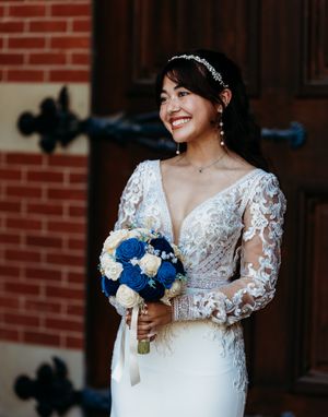 Smiling bride in lace long-sleeve gown holds blue and white rose bouquet by brick church doors.