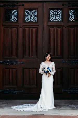 Elegant bride in lace long-sleeve gown with train poses before grand ornate wooden doors.