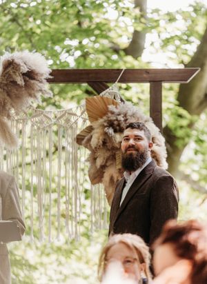 Bearded groom in brown tweed suit smiling at altar beneath boho macrame and pampas grass arch.
