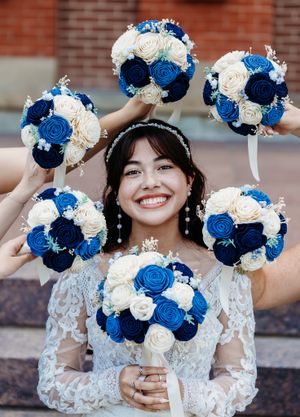 Radiant bride surrounded by bridesmaids holding matching blue and white rose bouquets on wedding day.
