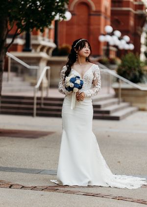 Bride in lace long-sleeve mermaid gown holds blue and white bouquet outside historic brick church.