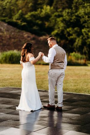 Bride in white off-shoulder gown and groom in vest hold hands dancing on outdoor tile floor at sunset.