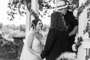 Black and white photo of smiling bride exchanging vows with officiant at outdoor wedding ceremony.