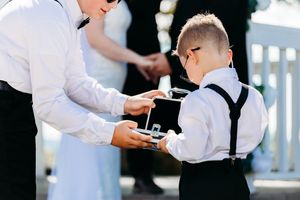 Young ring bearer in suspenders receiving wedding rings from older boy during outdoor ceremony.