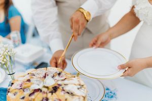 Close-up of couple cutting rustic berry wedding cake with gold utensils on elegant white gold-rimmed plate.