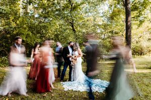 Bride and groom kissing surrounded by blurred wedding party guests in a lush outdoor garden setting.
