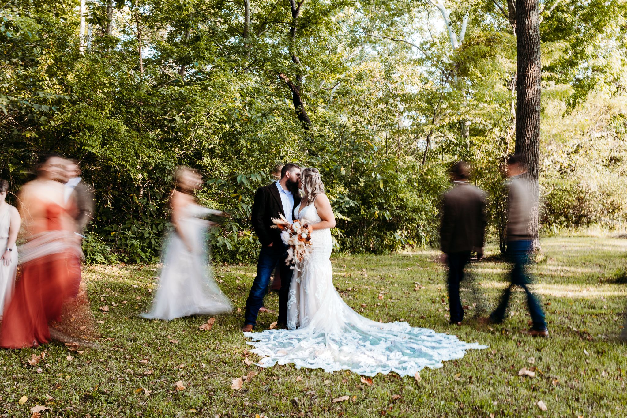 Wedding couple kissing while bridal party moves around them in a lush green outdoor ceremony setting.