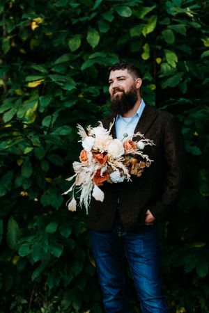 Bearded groom in brown blazer holding a boho bridal bouquet with orange and white flowers outdoors.