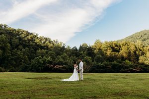 Bride and groom stand together in vast green meadow with wooded mountain backdrop under partly cloudy sky.