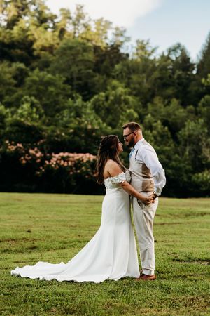 Bride in flowing white gown and groom in vest share romantic moment on lush green lawn.