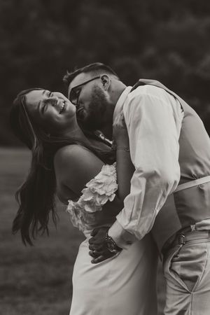 Black and white photo of laughing bride being kissed by bearded groom in sunglasses outdoors.