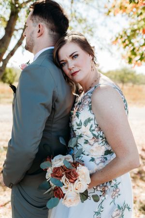 Bride in floral wedding dress leaning against groom holding a bouquet of rust and cream roses outdoors.