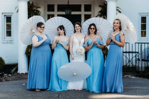 Bride and four bridesmaids in blue gowns pose holding white parasols outside white venue.