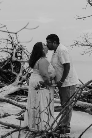 Black and white photo of couple kissing on beach surrounded by driftwood.