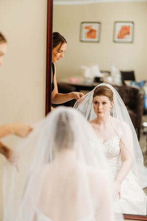 Bridesmaid carefully places veil on bride while she gazes into mirror during wedding preparations.