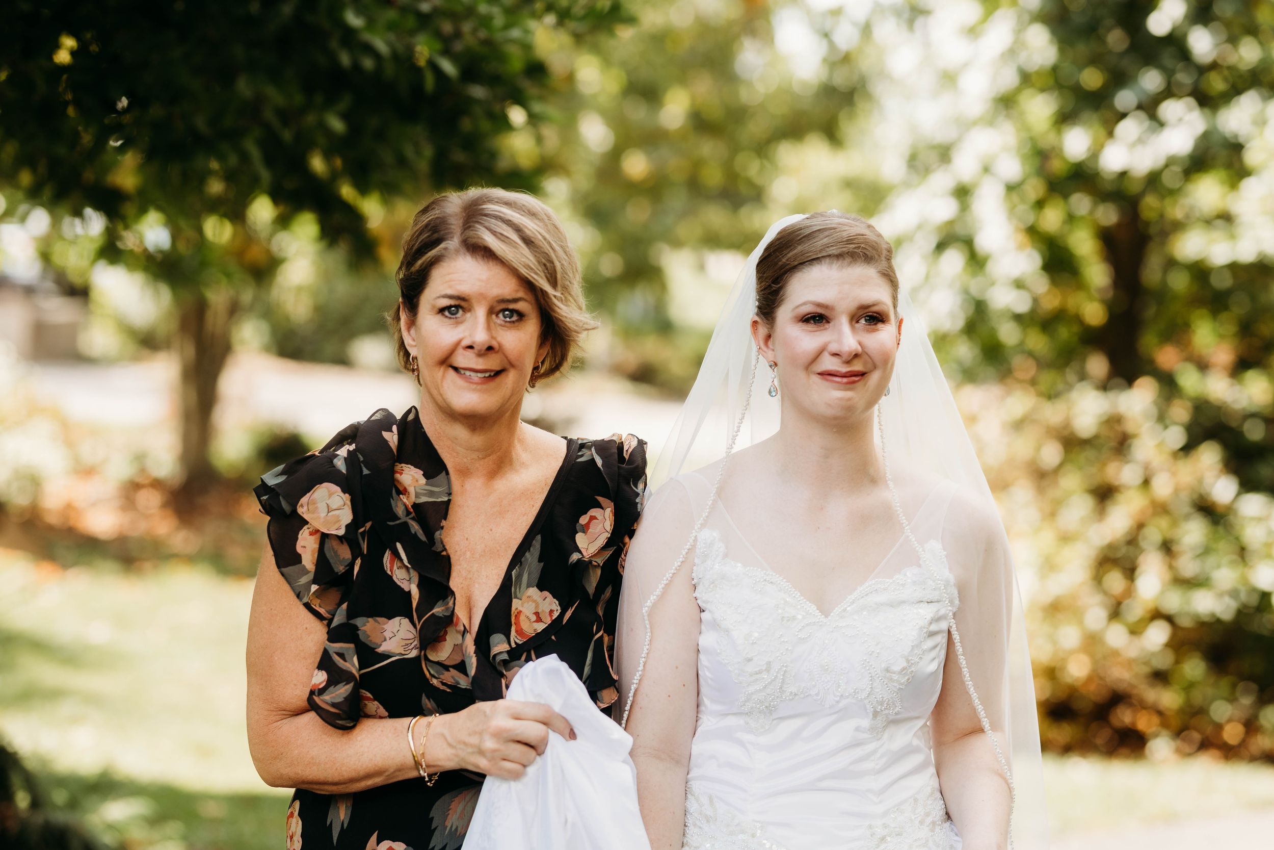 Bride poses with her mother outdoors before the wedding ceremony under lush green trees.